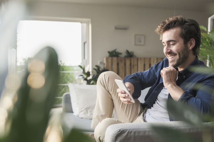 Man in a casual shirt and chinos smiling at a smartphone with plants surrounding him